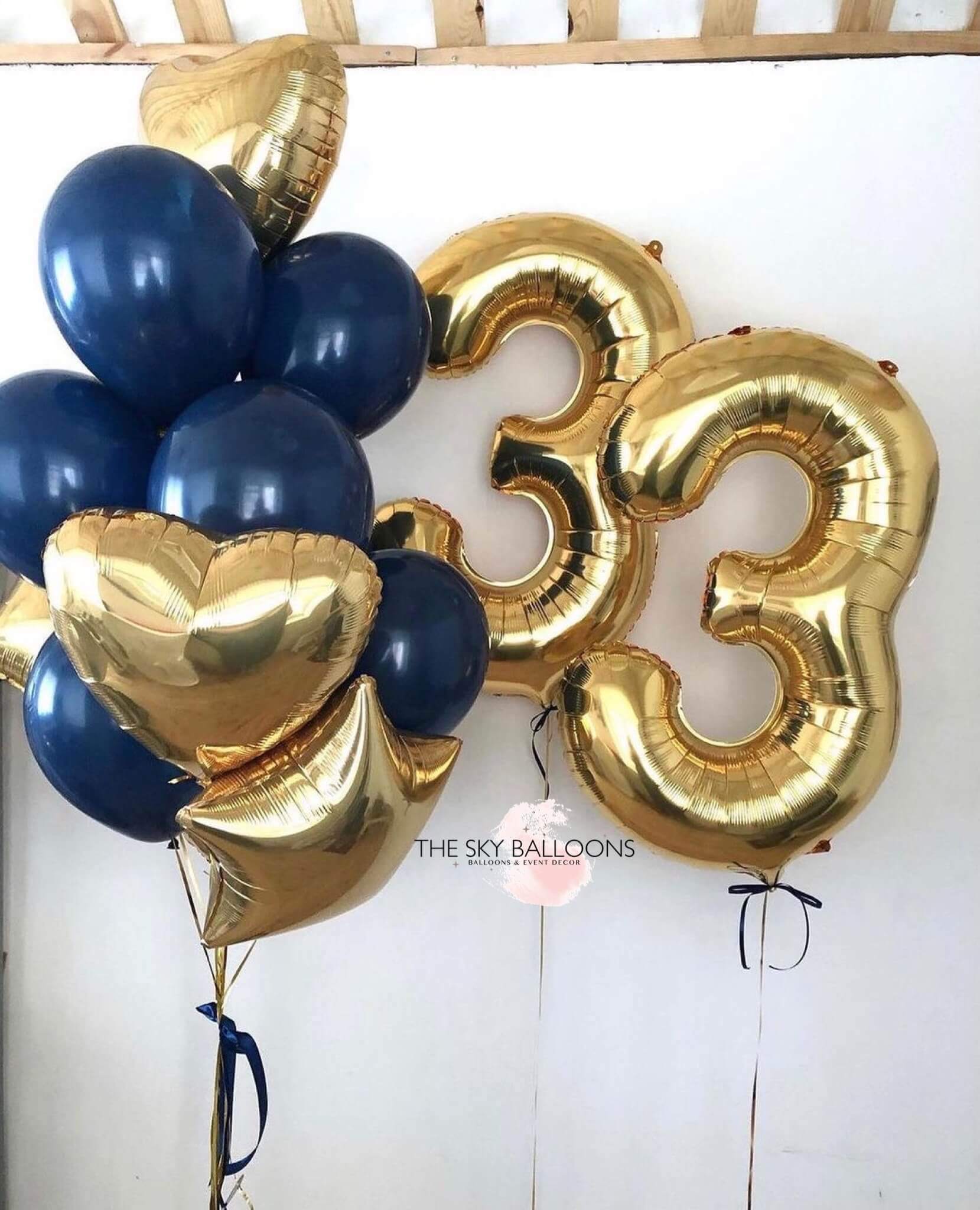 A bouquet of balloons including gold and navy blue colored latex balloons, with two gold star-shaped balloons, two gold heart-shaped balloons, and two gold numerical balloons spelling out a number.